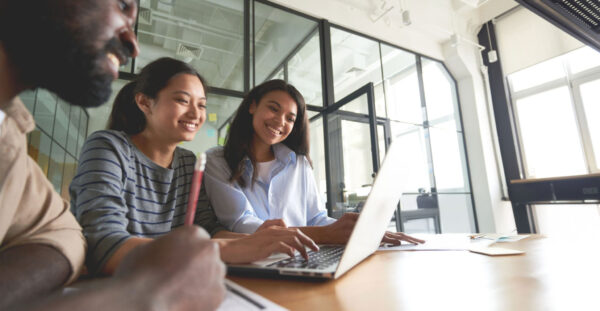 Copy space photo of two joyous women and smiley man looking at laptop screen and smiling while sitting at desk